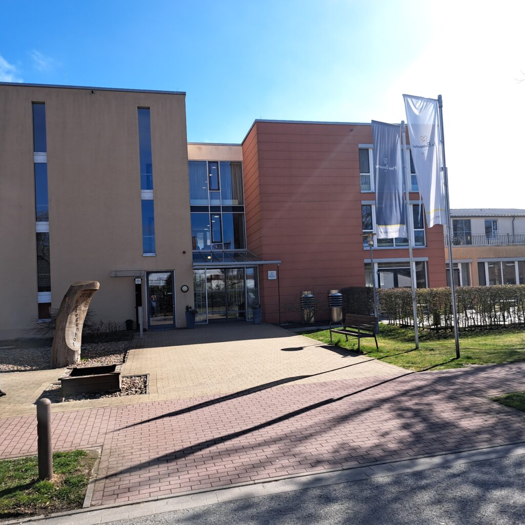 Frontansicht des Pflegeheims Haus Botegunhof Achim von Korian: moderner Haupteingang mit Glasvordach, Fahnen und gepflegter Außenanlage an einem sonnigen Tag