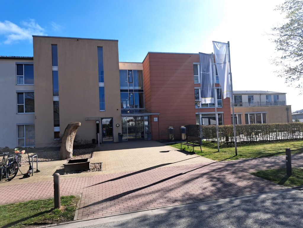 Frontansicht des Pflegeheims Haus Botegunhof Achim von Korian: moderner Haupteingang mit Glasvordach, Fahnen und gepflegter Außenanlage an einem sonnigen Tag