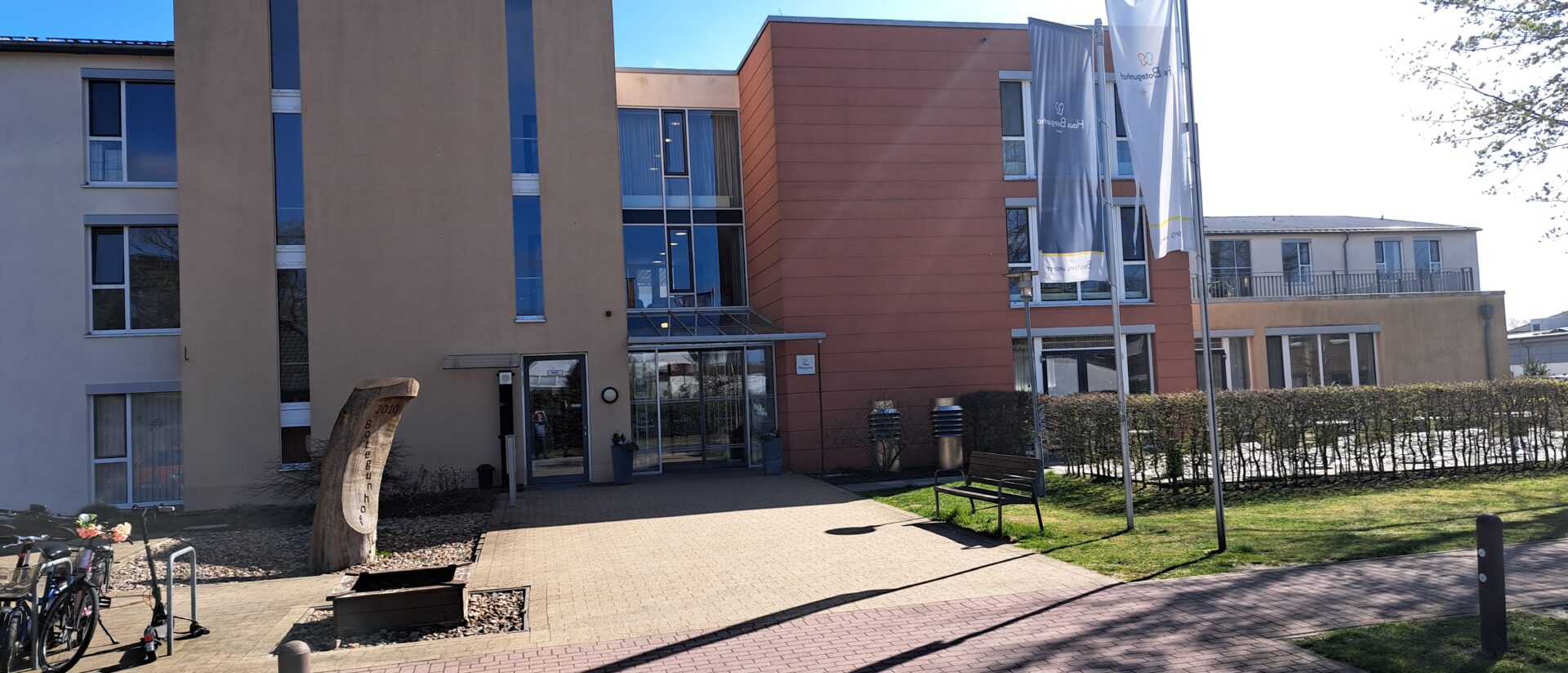 Frontansicht des Pflegeheims Haus Botegunhof Achim von Korian: moderner Haupteingang mit Glasvordach, Fahnen und gepflegter Außenanlage an einem sonnigen Tag
