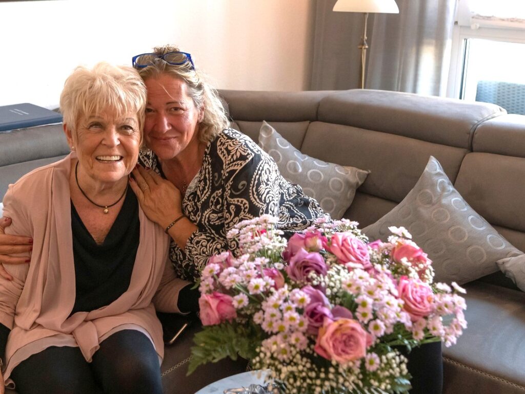 Pflegekraft umarmt lachende Seniorin auf Couch mit Blumen beim ambulanten Pflegedienst Stadtpark Hilden