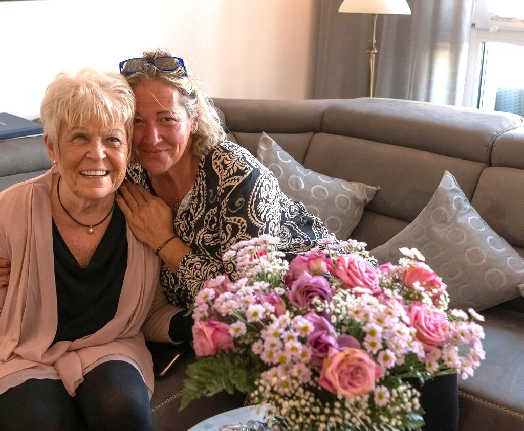 Pflegekraft umarmt lachende Seniorin auf Couch mit Blumen beim ambulanten Pflegedienst Stadtpark Hilden