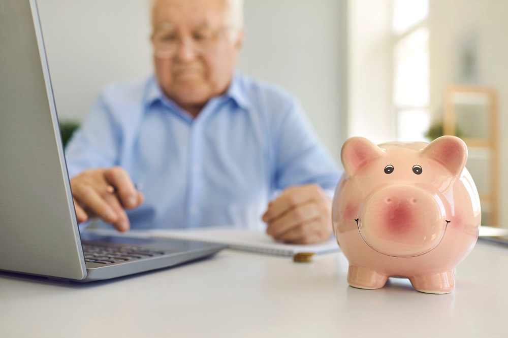 Pflegegeld Piggy bank standing on desk with blurred senior man using laptop to pay bills online in background
