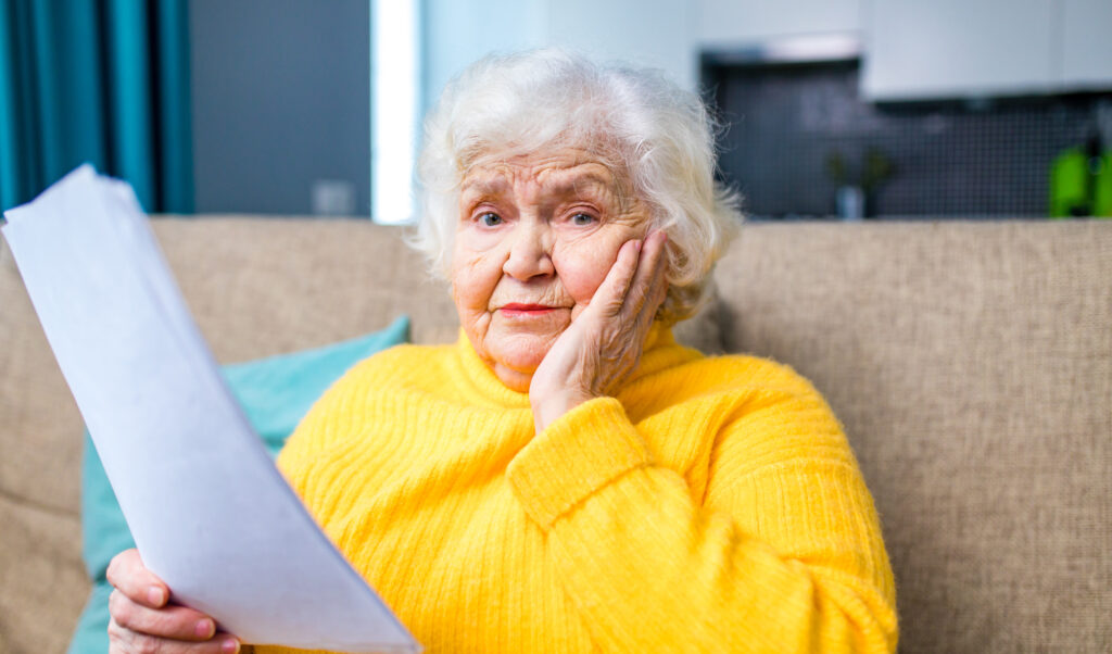 Hilfe zur Pflege shoked amazed old woman white gray-haired sitting on the sofa in living room with laptop and bills