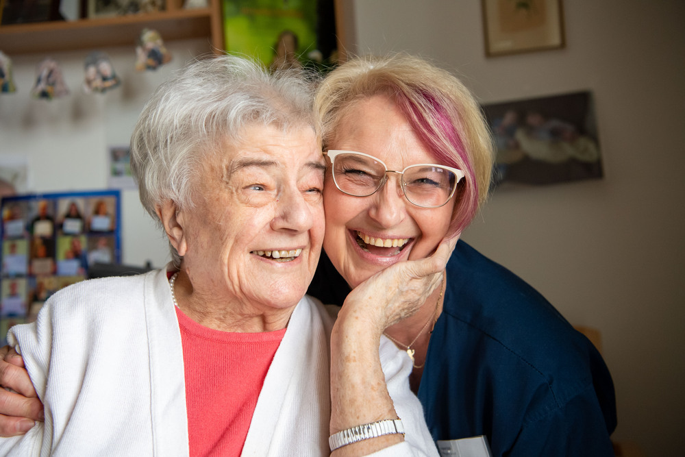 Nahaufnahme Seniorin und Pflegerin im Pflegeheimzimmer der Seniorin. Beide lachen.