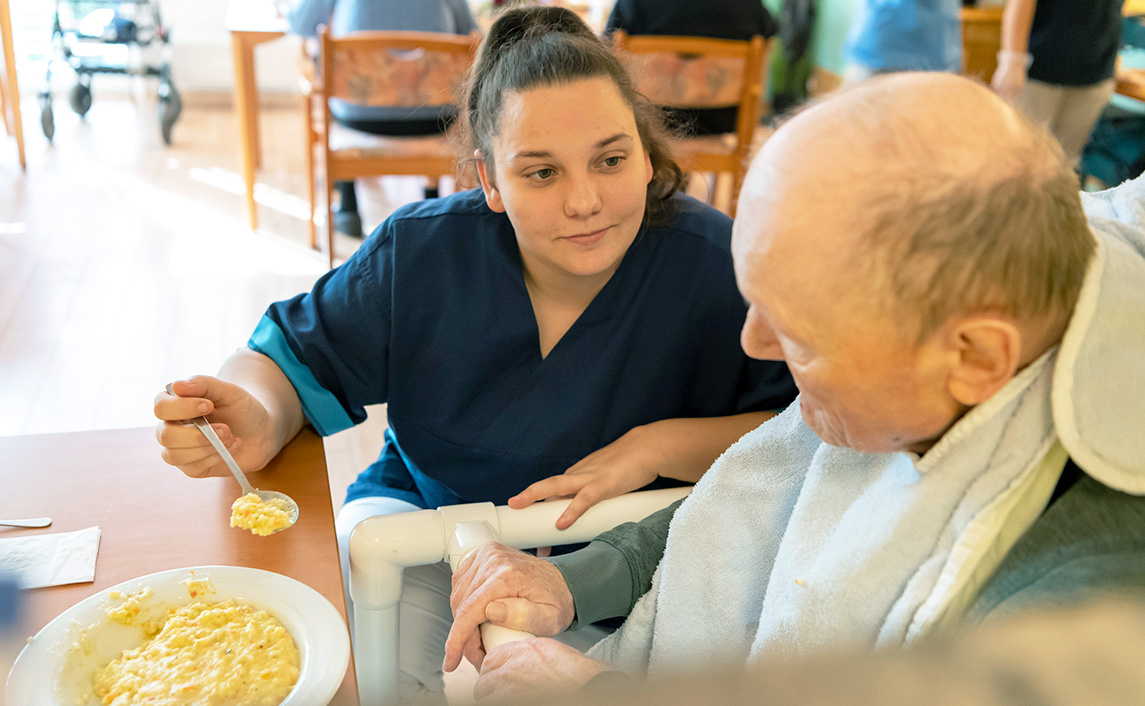Pflegekraft unterstützt älteren Mann beim Essen im Pflegeheim, barrierefreies Essen bei Schluckstörungen