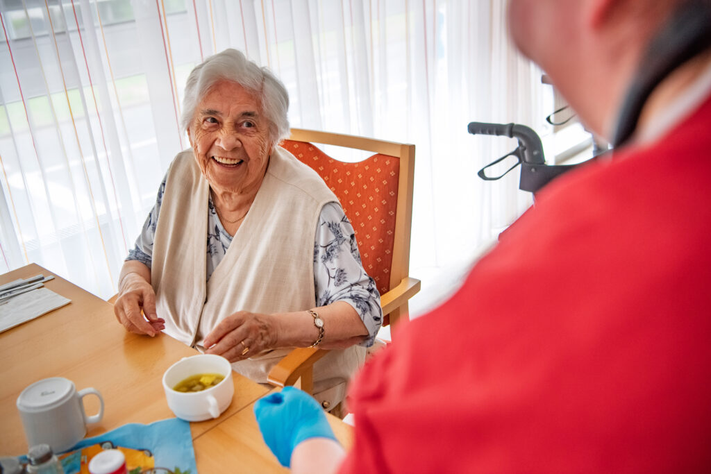 Zukunftspakt Pflege, Ältere Dame sitzt am Tisch mit Kaffee und lacht, im Vordergrund eine Person mit rotem Oberteil