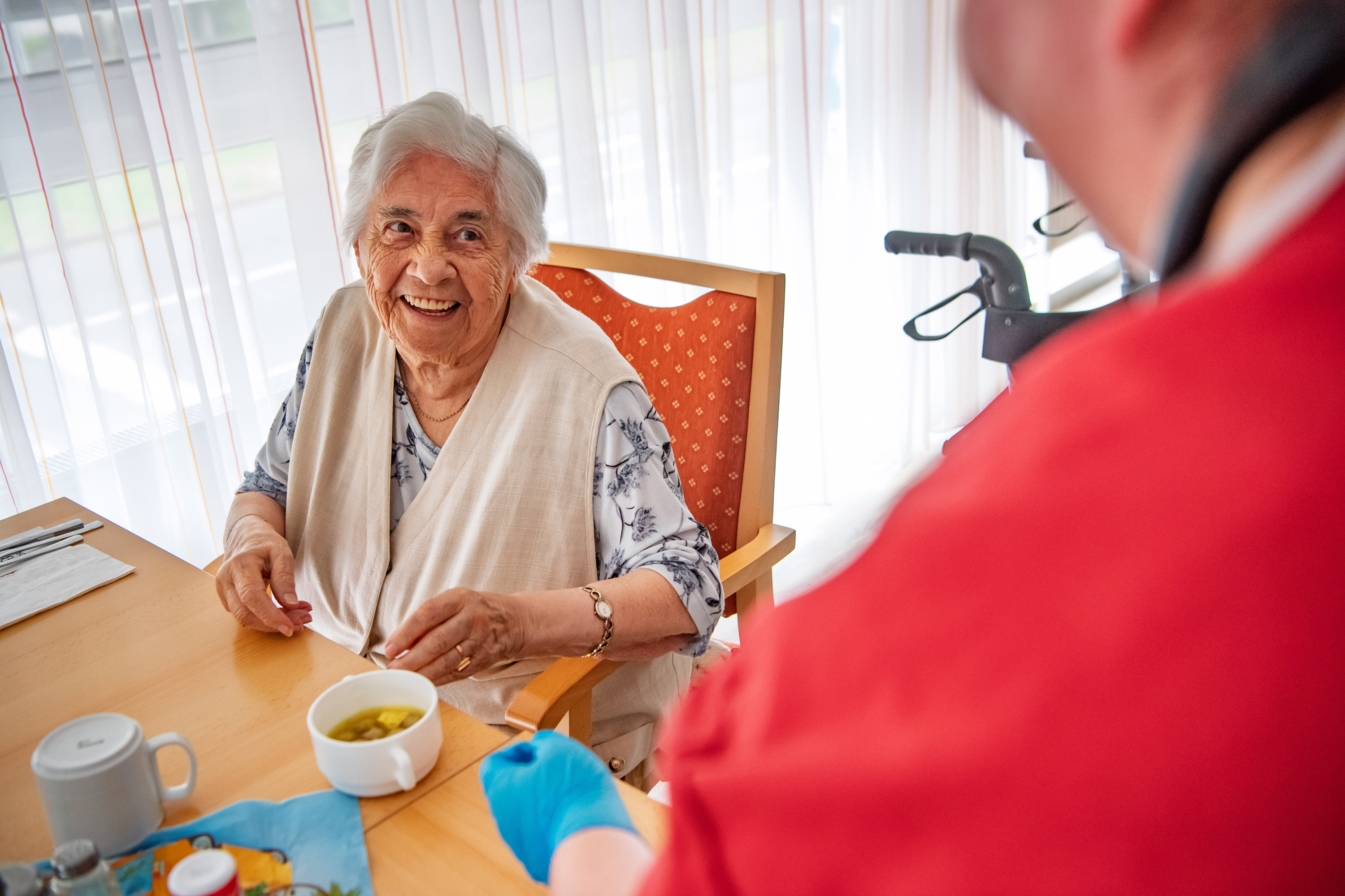 Zukunftspakt Pflege, Ältere Dame sitzt am Tisch mit Kaffee und lacht, im Vordergrund eine Person mit rotem Oberteil