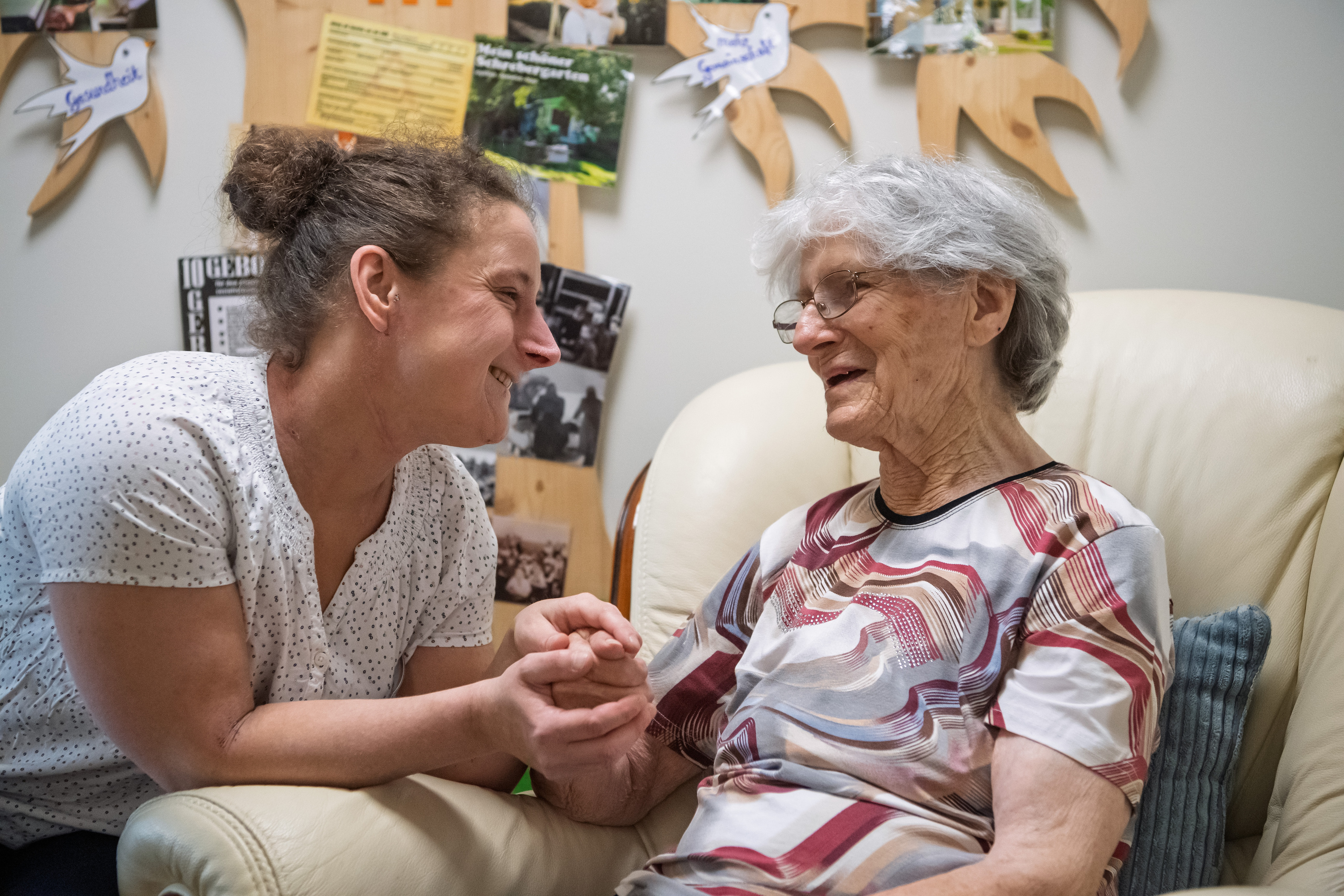 Diskussion um Pflegegrad 1 - Seniorin sitzt in Sessel, ihr gegenüber sitzt eine jüngere Frau und beugt sich zu ihr, beide lachen