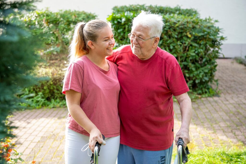 Pflegekraft begleitet Senior mit Rollator im Garten einer Pflegeeinrichtung