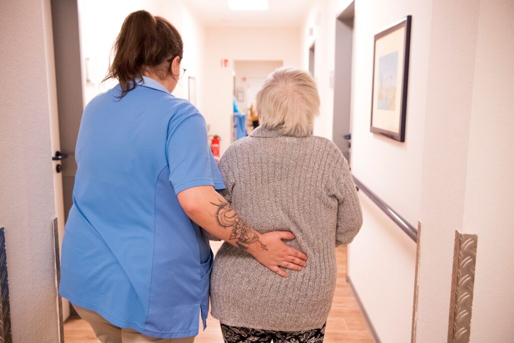 Weibliche Pflegefachkraft von Korian begleitet Senior in auf dem Flur eines Pflegeheims von hinten – Symbolbild für steigende Pflegekosten in Deutschland