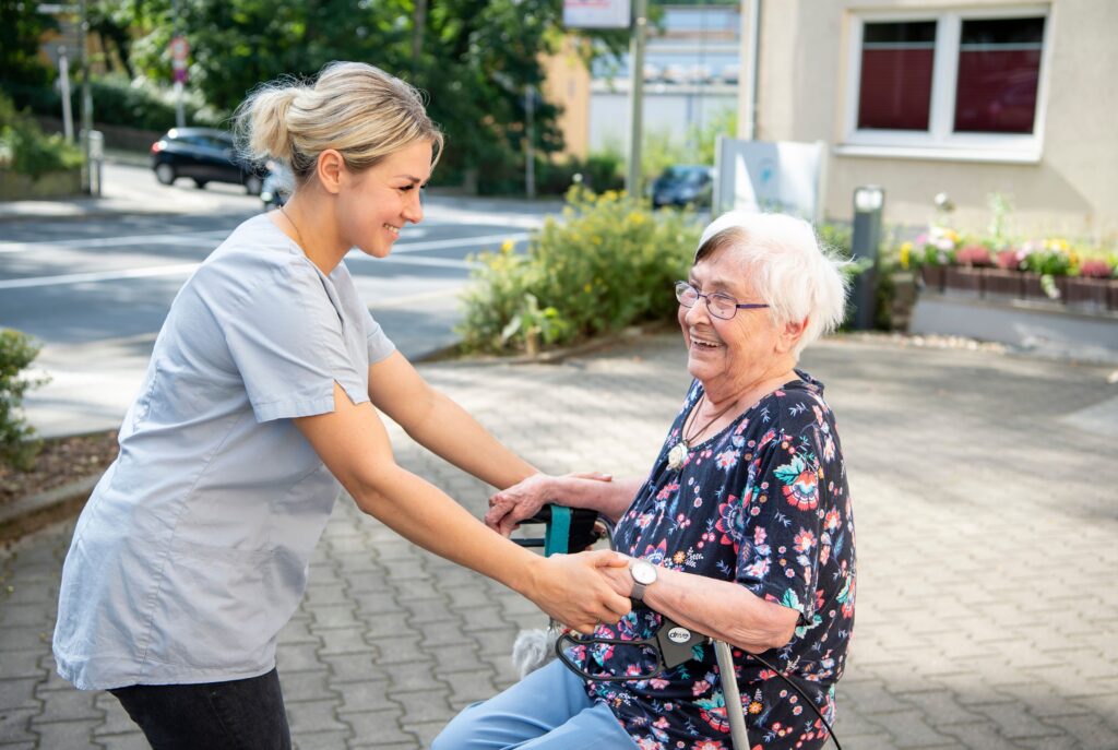 Pflegekraft hilft Seniorin beim Aufstehen mit Rollator nach Oberschenkelhalsbruch